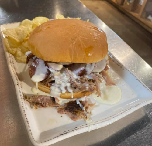A pulled pork sandwich with creamy sauce, served with a side of potato chips on a white plate.