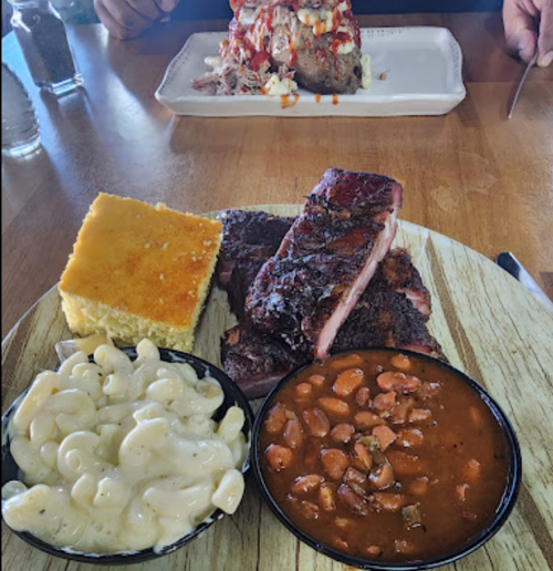 A plate of barbecue ribs, macaroni and cheese, baked beans, and cornbread on a wooden table.