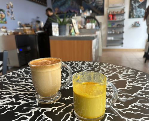 Two drinks on a patterned table: a latte in a clear mug and a golden turmeric drink in a yellow glass, with a café background.