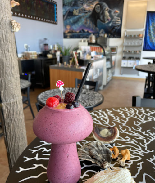 A colorful smoothie in a mushroom-shaped glass, garnished with berries, sits on a patterned table in a cozy café.