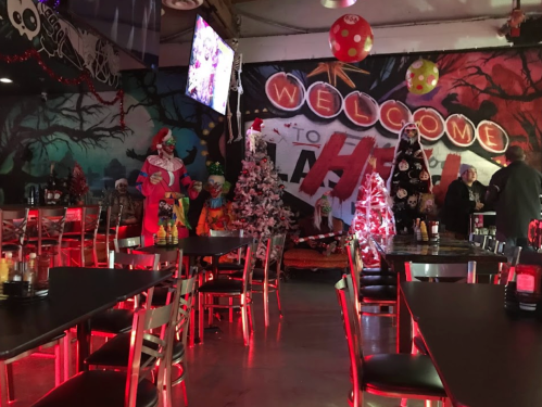 A festive restaurant interior with clown decorations, Christmas trees, and a colorful mural, featuring red lighting and tables.