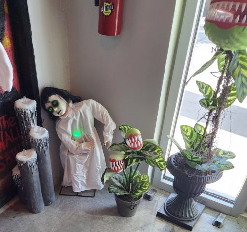 A spooky scene featuring a ghostly doll, eerie plants, and candles in a corner by a window.