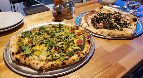 Two pizzas on metal trays: one topped with greens and tomatoes, the other with greens and meat, on a wooden table.