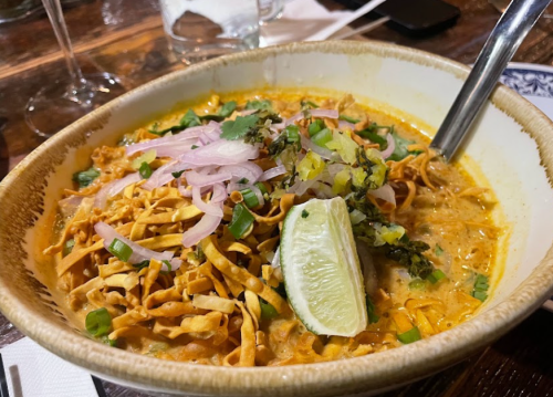 A bowl of creamy noodle soup topped with crispy noodles, lime, onions, and herbs on a wooden table.