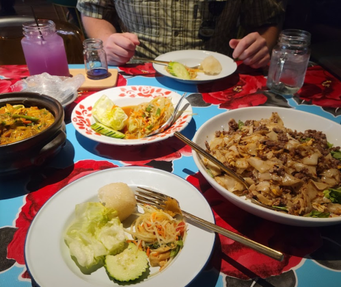 A table with various dishes: a large bowl of stir-fried noodles, salad, and a clay pot dish, with drinks in the background.