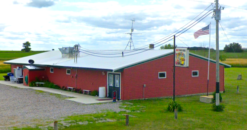 A red building with a white roof, featuring a sign and an American flag, set in a rural landscape with fields.