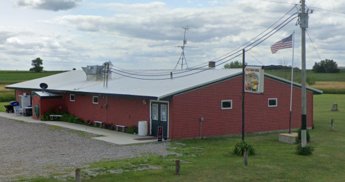 A red building with a white roof, surrounded by fields, featuring a sign and an American flag on a pole.