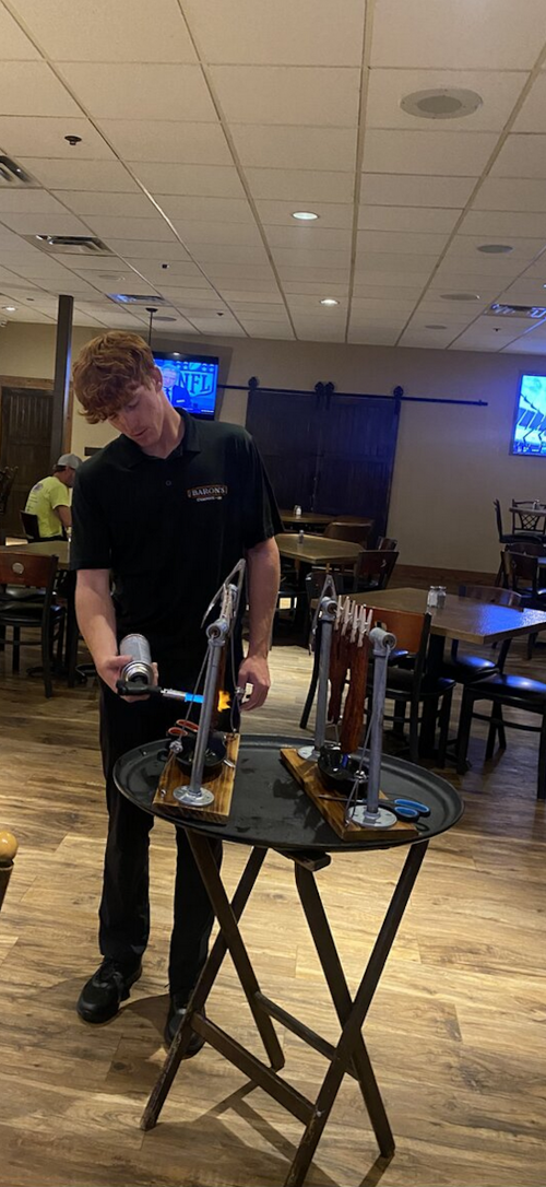 A server in a black shirt uses a torch on a wooden display in a restaurant setting. Tables and TVs are in the background.