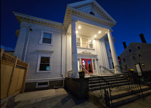 A grand white building with columns, illuminated at night, featuring a red door and a staircase leading up to it.