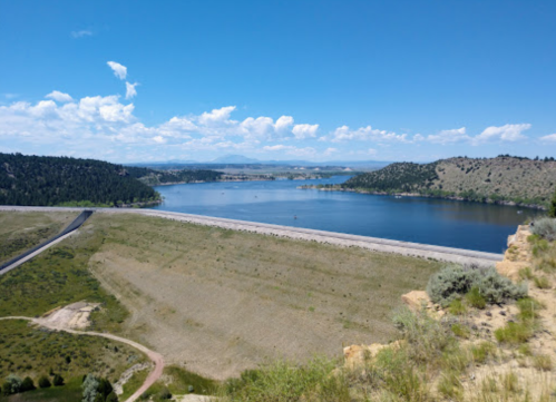 A scenic view of a lake surrounded by hills and greenery under a clear blue sky with scattered clouds.