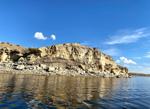 A rocky shoreline under a clear blue sky, reflecting in calm water with scattered clouds above.