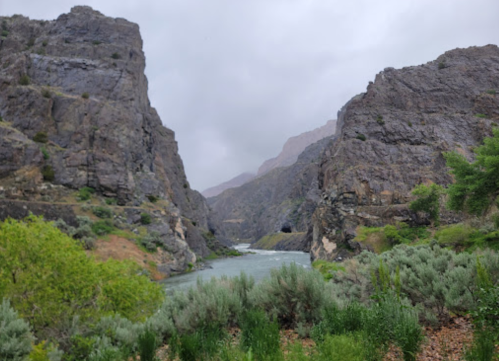 A river flows between steep rocky cliffs, surrounded by greenery under a cloudy sky.