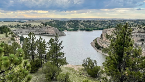 A serene river winding through green hills and rocky cliffs under a cloudy sky. Pine trees frame the view.