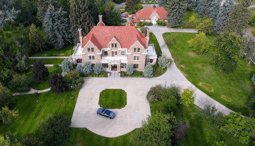 Aerial view of a large, elegant mansion surrounded by lush greenery and a circular driveway.