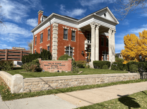 Historic brick building with columns, surrounded by greenery and autumn trees, labeled as a state office in Wyoming.