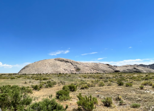 A large, smooth rock formation rises above a grassy landscape under a clear blue sky.