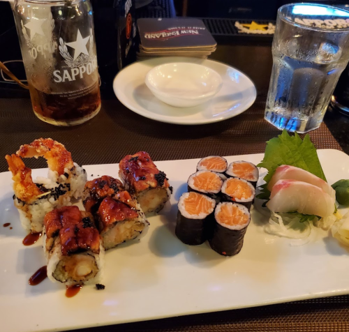 A plate of sushi featuring various rolls, sashimi, and garnishes, with a drink and a small dish in the background.