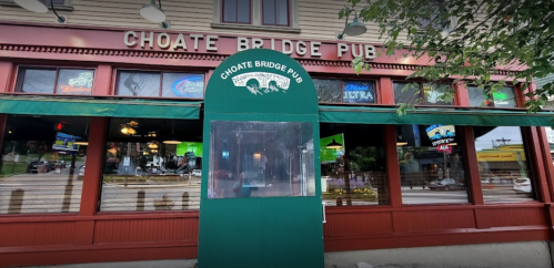 Exterior of Choate Bridge Pub featuring a green entrance and large windows with various signs and outdoor seating.