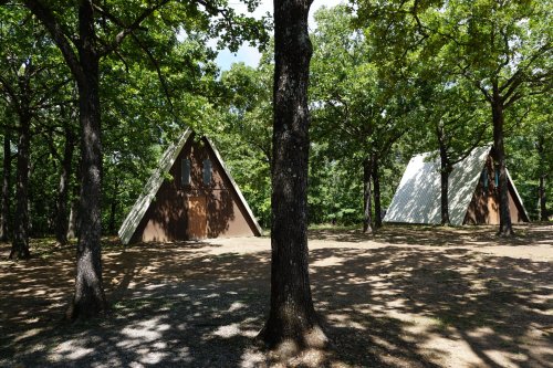 Two A-frame cabins nestled among trees in a wooded area, with dappled sunlight filtering through the leaves.