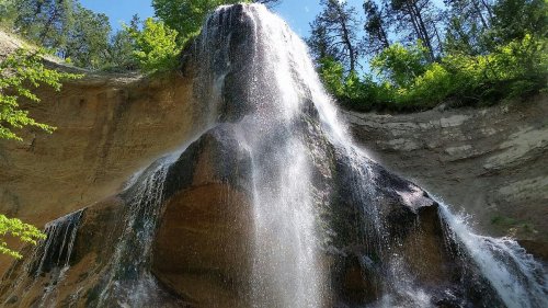 A cascading waterfall flows down a rocky cliff, surrounded by lush green trees under a clear blue sky.