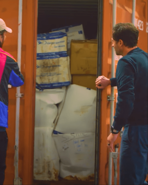 Two men stand by an open shipping container filled with stacked boxes and bags.