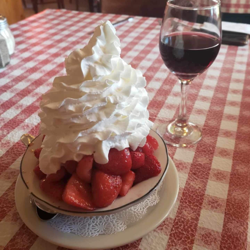 A bowl of strawberries topped with whipped cream, alongside a glass of red wine on a checkered tablecloth.