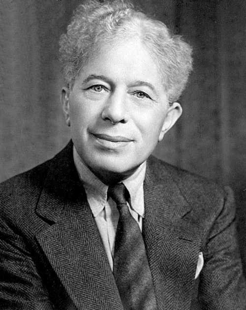 Black and white portrait of a man with curly hair, wearing a suit and tie, smiling at the camera.