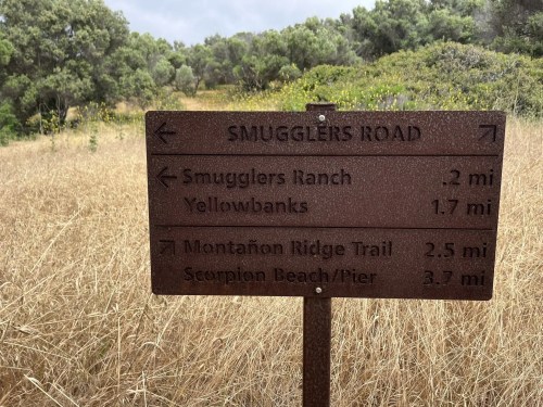A brown directional sign indicating distances to Smugglers Ranch, Yellowbanks, Montañon Ridge Trail, and Scorpion Beach/Pier.