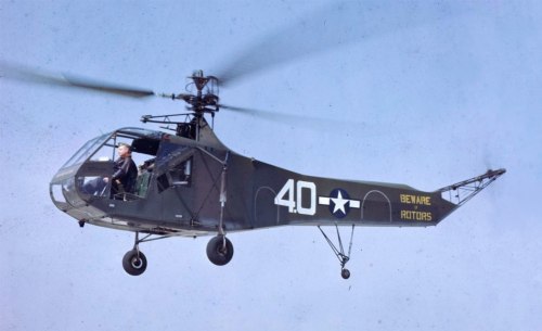 A military helicopter in flight, marked with "40" and "Beware Rotors," with a pilot visible in the cockpit.