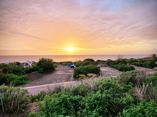 Sunset over a coastal campsite, with tents and lush greenery in the foreground and a serene ocean view.