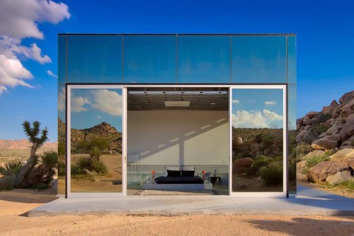 Modern glass house in a desert landscape, reflecting surrounding rocks and plants under a blue sky with clouds.