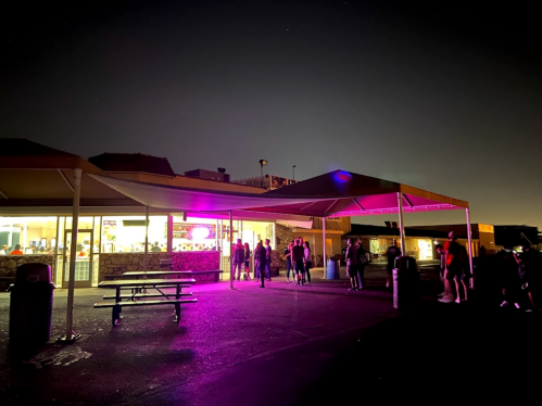 A brightly lit building at night with people gathered outside under a canopy, illuminated by purple lights.
