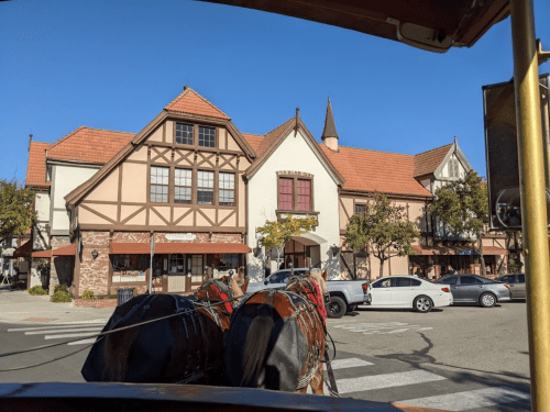 A view of a charming town square with horses in front, featuring European-style buildings and clear blue skies.