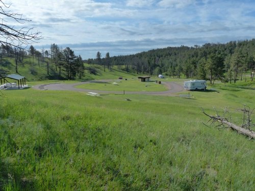 A scenic view of a grassy campground with trees, a winding road, and a few parked RVs under a cloudy sky.