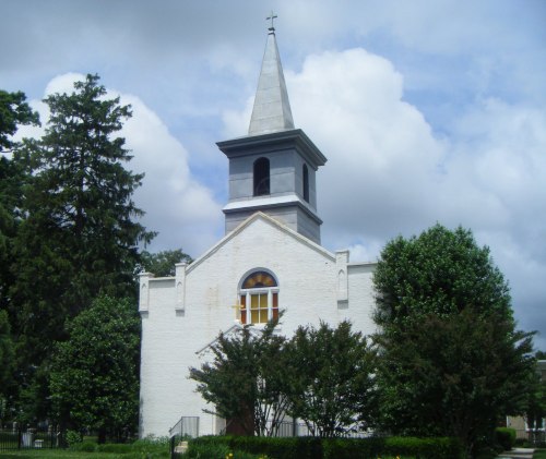 A white church with a tall steeple, surrounded by trees and under a cloudy sky.