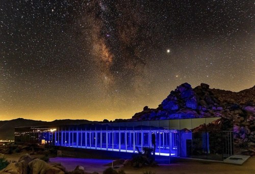 A modern glass building illuminated in blue under a starry night sky with the Milky Way visible above.