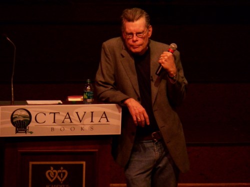 A speaker in a gray blazer leans against a podium with a microphone, looking thoughtful at a book event.