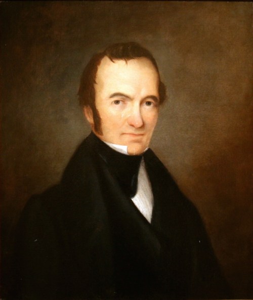 Portrait of a man in a dark coat with a white collar, set against a muted brown background.