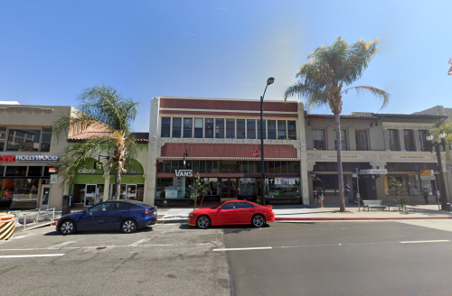 A street view featuring shops, including Vans and Olive, with palm trees and parked cars under a clear blue sky.