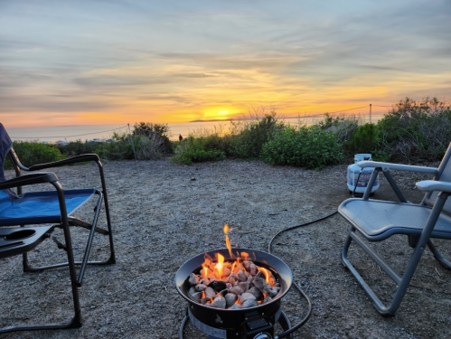 A cozy campsite with two chairs facing a fire pit, set against a vibrant sunset over the ocean.