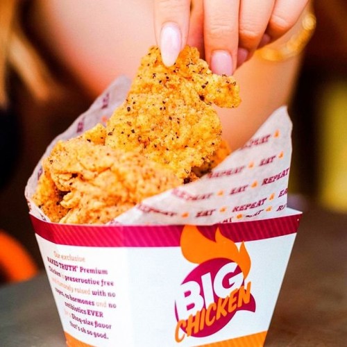 A hand reaching into a box of crispy fried chicken pieces with a colorful logo on the container.
