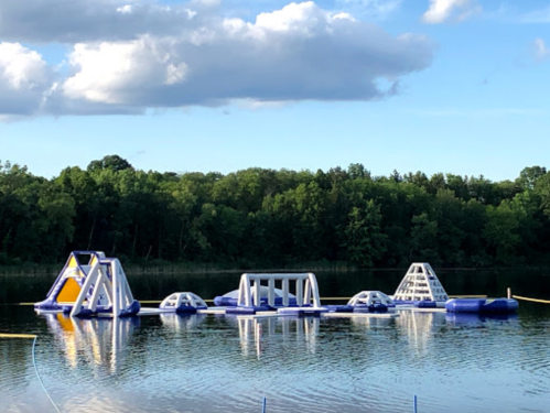 Inflatable water obstacle course on a lake, surrounded by trees and under a partly cloudy sky.