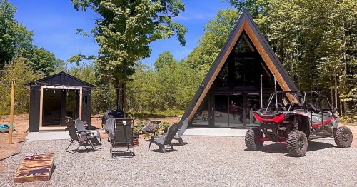 A modern A-frame cabin surrounded by trees, with outdoor seating and an off-road vehicle parked nearby.