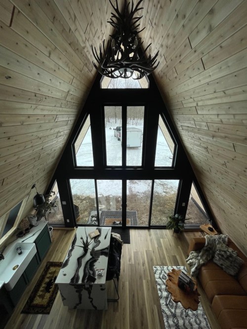 A cozy A-frame cabin interior with wooden walls, large windows, and a view of snow-covered ground outside.