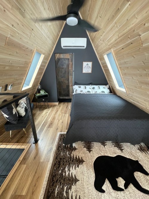 Cozy A-frame interior with wooden walls, a black bedspread, bear-themed decor, and a ceiling fan.