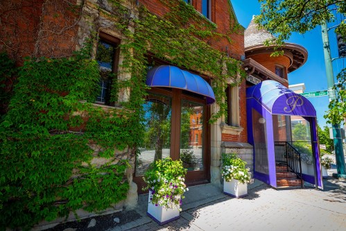 A charming brick building with blue awnings, surrounded by greenery and flower planters, on a sunny day.