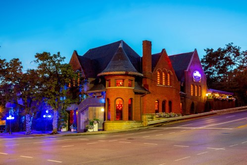 A charming brick building with a steep roof, illuminated by colorful lights, set against a twilight sky.