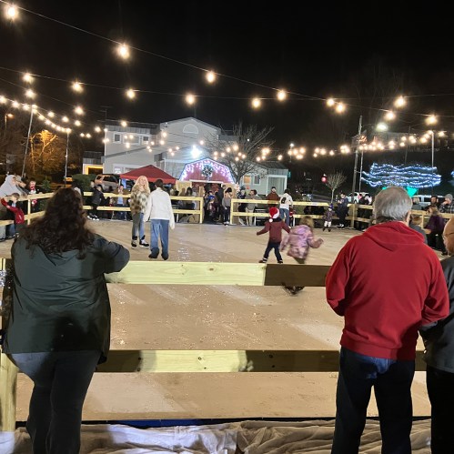 A festive outdoor ice skating rink with people skating, surrounded by string lights and a crowd watching.
