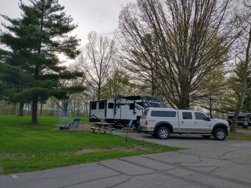 A camper and truck parked in a grassy area surrounded by trees, with a picnic table and fire pit nearby.