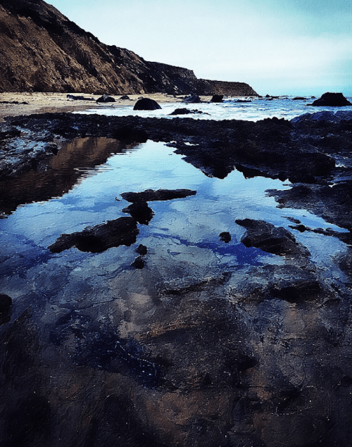 A rocky shoreline with calm water reflecting the sky, surrounded by cliffs and a serene beach landscape.